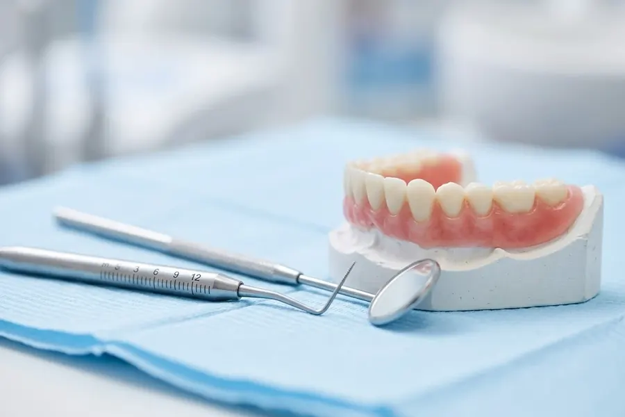 A periodontal probe and dental mirror on a blue cloth beside a dental model with pink gums.